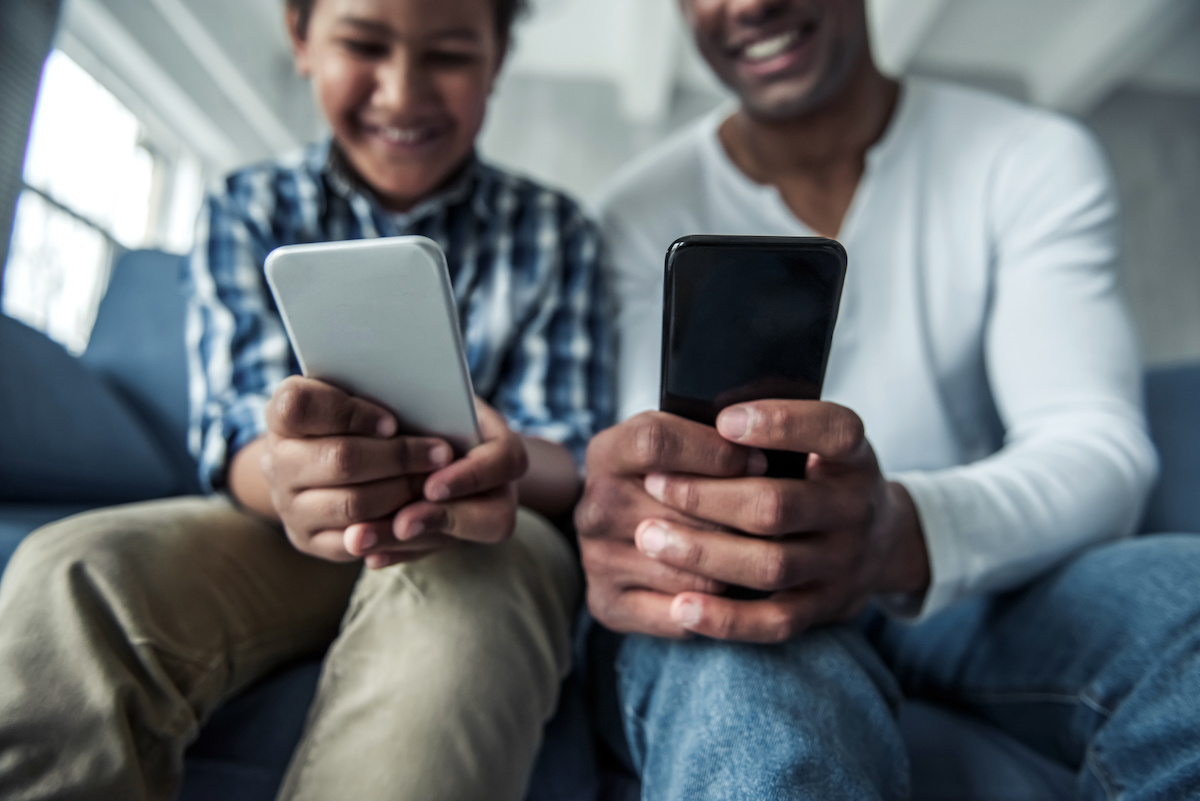 Cropped image of father and son in casual clothes using smartphones and smiling while sitting on couch at home
