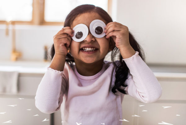 Child holding large googlie eyes in front of her eyes and smiling.