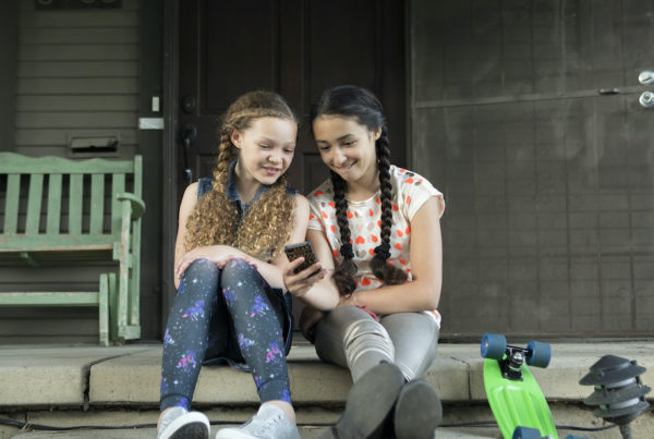 Two young teens sitting next to one another on a porch and looking at a smartphone.