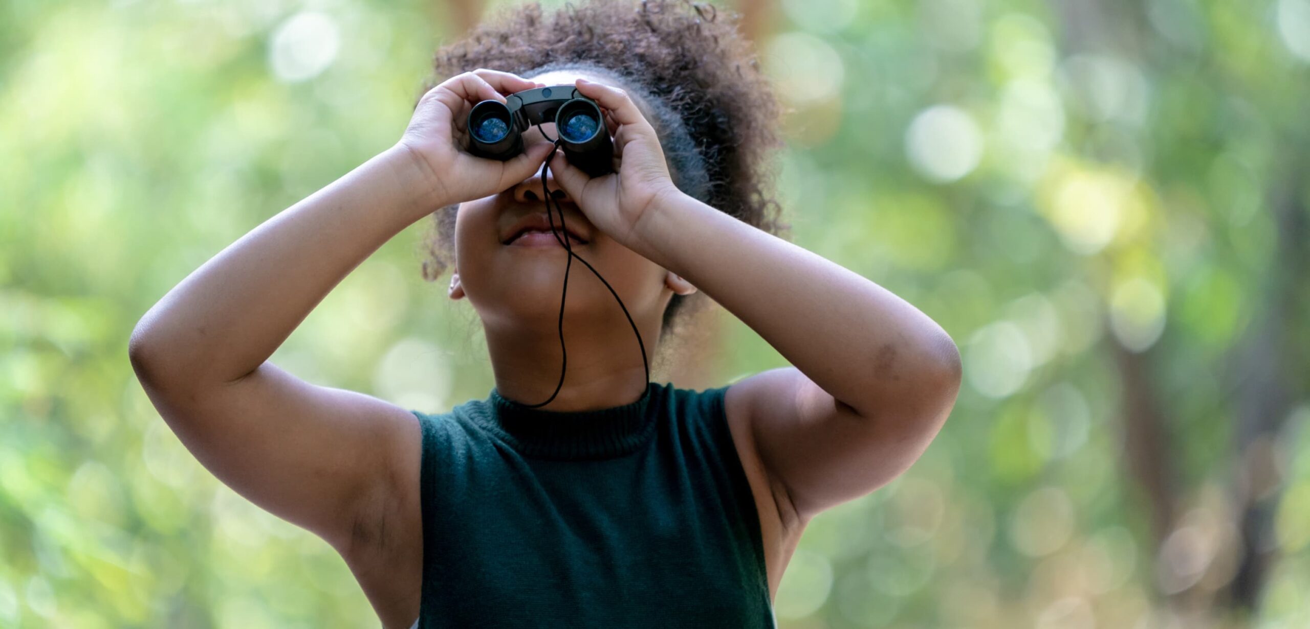 A young girl peers up into the canopy above, with a pair of bird watching binoculars.